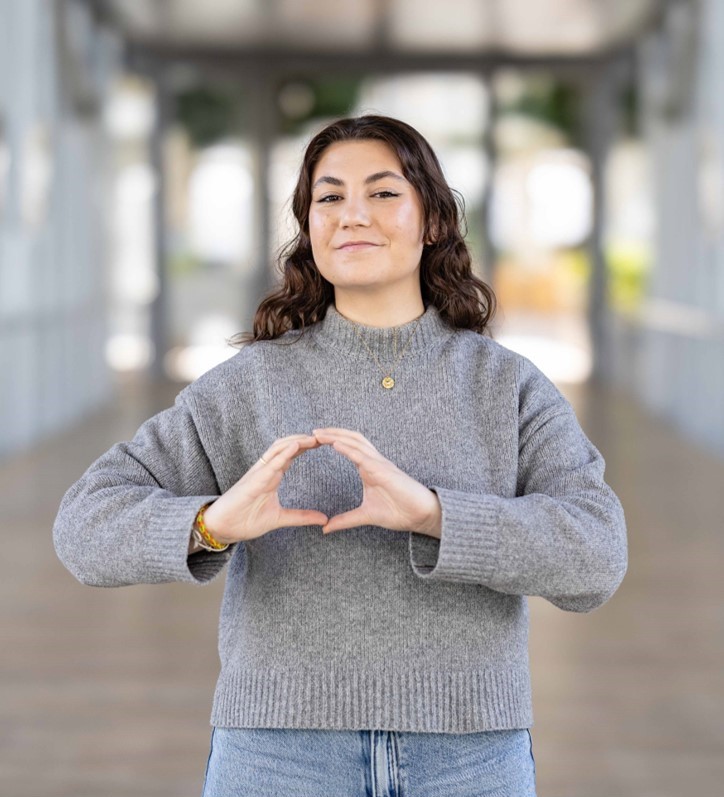 Femme souriant, les mains formant le logo One Health, portant un pull gris et un jean, devant un bâtiment moderne.