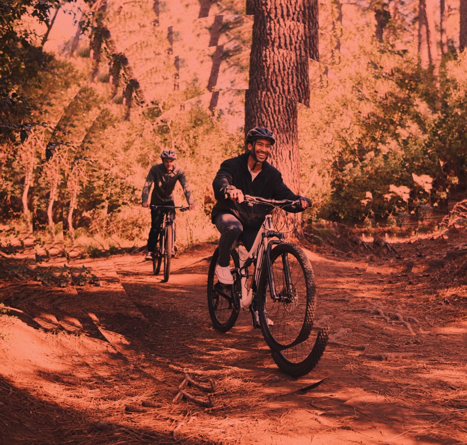 Deux cyclistes descendent une piste forestière en terre, entourés d'arbres. L'avant-plan montre un cycliste souriant, portant un casque et des vêtements de sport. Un autre cycliste suit derrière.